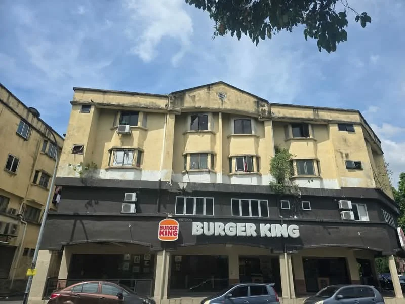 Exterior type: low-rise apartment block; features: yellow painted facade, multiple air conditioning units visible, ground floor commercial space with signage, second floor with windows, overgrown plants on the facade, two-storey structure, flat roof, urban setting with street view; style: traditional.