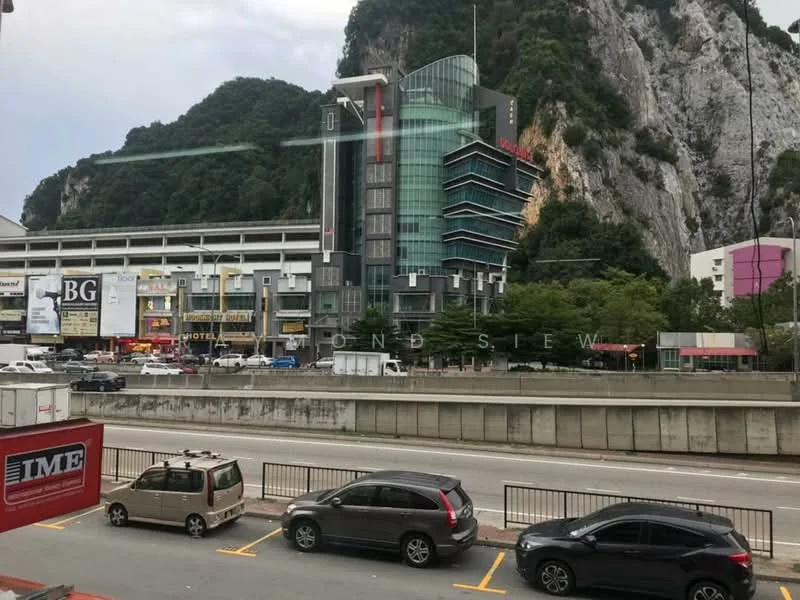 Exterior type: mixed-use development; features: glass curtain wall facade, multi-storey building, surrounded by mountains, commercial spaces on ground floor, parking area in front, urban street view, visible signage for businesses, landscaped area with trees; style: modern.
