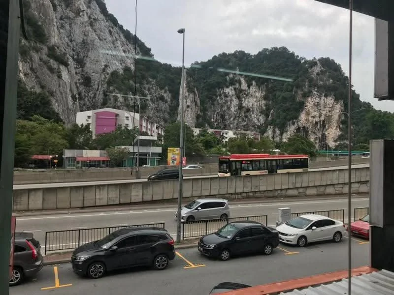 Exterior features: view of rocky hills, road with vehicles, urban setting, trees along the roadside, commercial buildings in the background, bus on the road, parking spaces visible.