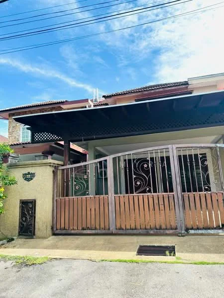 Exterior type: semi-detached house; features: metal gate with decorative design, cream painted facade, wooden balcony with lattice design, single-storey, concrete pathway, manicured garden area, surrounded by greenery, sloped roof, visible utility lines; style: traditional.