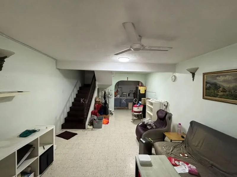Living features: light-colored painted walls, tile flooring, 5-blade ceiling fan, recessed ceiling light, sofa in grey, coffee table, wall-mounted artwork, open shelving unit, area rug, staircase leading to upper floor. Kitchen features: kitchen area visible in background, white cabinetry, sink, appliances visible, laundry area with washing machine.