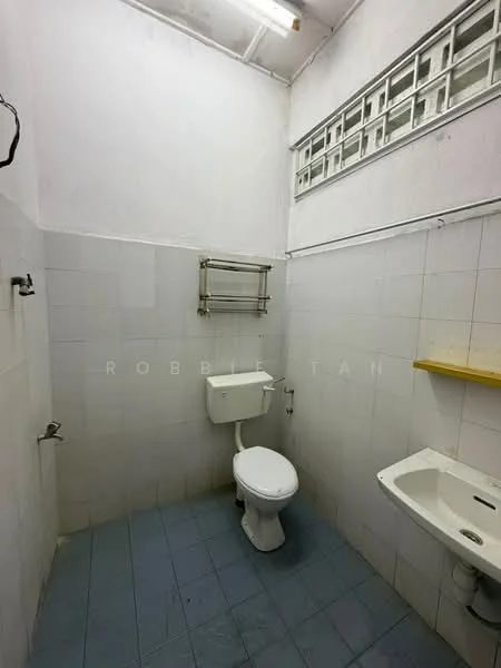 Bathroom features: white tiled walls, blue ceramic tile flooring, one-piece toilet, wall-mounted sink, metal towel rack, single wall-mounted shelf, fluorescent ceiling light, frosted glass block window.