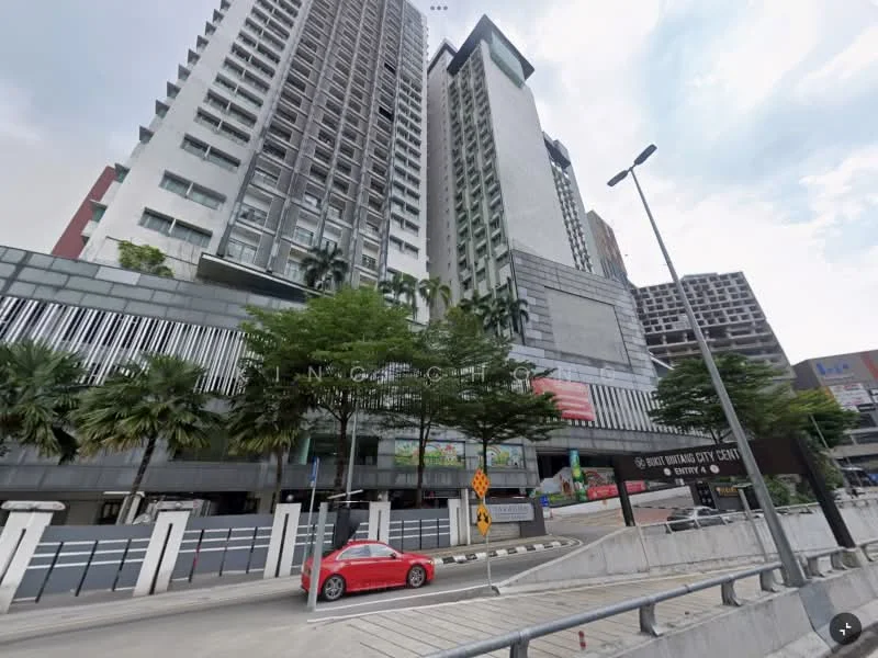 Exterior type: high-rise condominium; features: glass curtain wall, exposed concrete facade, recessed balconies, vertical fins, manicured landscaping with palm trees, surrounded by mature greenery, driveway with interlocking pavers, signage visible on the building, multiple storeys, urban context with nearby buildings; style: modern.