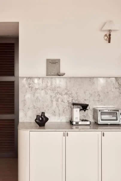 Kitchen features: white cabinetry, marble countertop, marble backsplash, built-in coffee machine, toaster, wall-mounted sconce, dark ceramic vase, neutral wall color, sliding door, wooden louvered panels; style: modern.