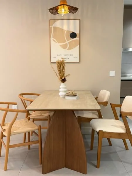 Dining features: wooden dining table with unique base, 4 dining chairs with wooden frames, upholstered seat on two chairs, pendant light fixture, framed artwork on wall, neutral painted walls, decorative vase with dried flowers, table setting with book and decorative item; style: modern.