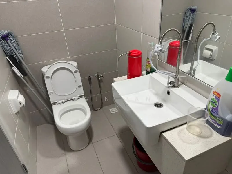 Bathroom features: white toilet, single-sink vanity with stone countertop, chrome basin mixer faucet, wall-mounted mirror, grey tiled walls, grey tiled flooring, toilet paper holder, cleaning supplies visible, glass tumbler, red bottle, red container; style: modern.
