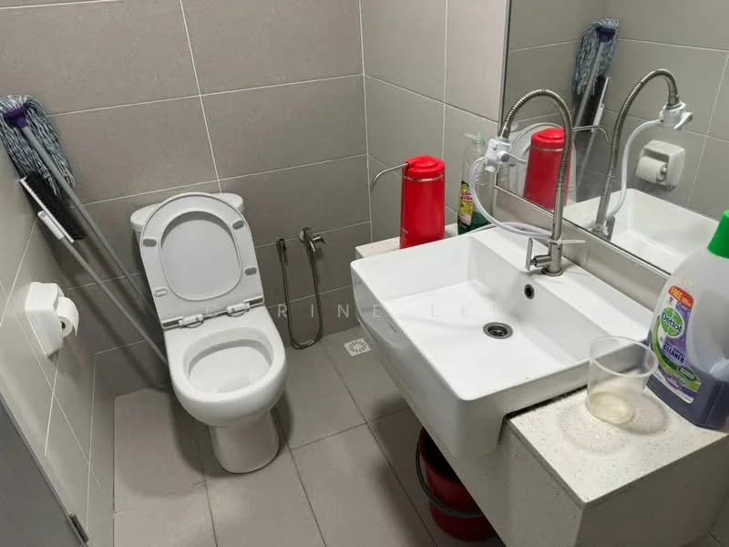 Bathroom features: white toilet, single-sink vanity with stone countertop, chrome basin mixer faucet, mirror above sink, grey tiled walls, light grey tiled flooring, toilet paper holder, cleaning supplies visible, glass cup on countertop, red bottle on countertop; style: modern.