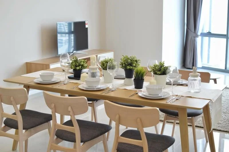 Dining features: 6-seater dining table, wooden dining table with beige table runner, white ceramic plates, glass wine glasses, decorative potted plants, cutlery set, beige upholstered dining chairs, TV console with wall-mounted TV, light-colored wall, floor-to-ceiling window with sheer curtains, area rug, neutral-colored walls; style: modern; view: city.