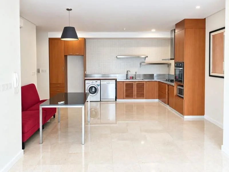 Kitchen features: brown wooden cabinetry, stainless-steel sink, chrome pull-down faucet, built-in oven, freestanding range oven, stainless-steel refrigerator, washing machine, grey tiled backsplash, marble tile flooring, recessed downlights, pendant light, table with metal legs, red fabric sofa; style: modern.