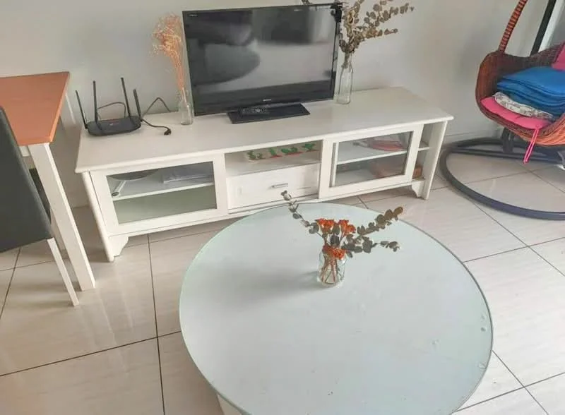 Living features: white TV console with glass doors, flat-screen TV, decorative vase with dried flowers, round glass coffee table, black router, dining table with wooden top, green dining chair, hanging chair, stack of folded blankets, white tiled flooring; style: modern.
