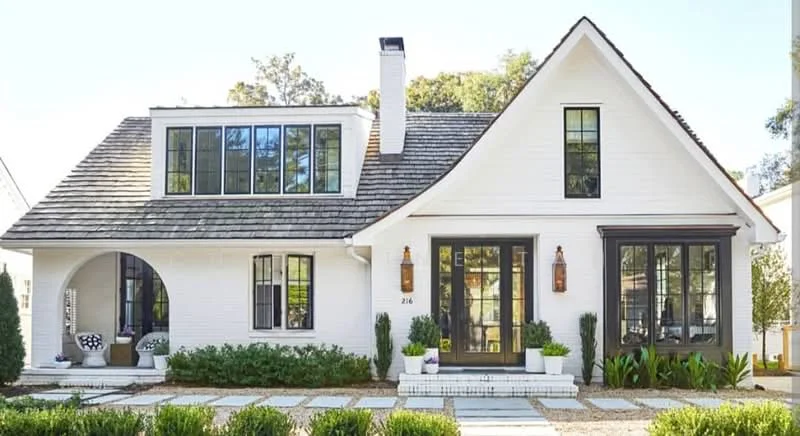 Exterior type: detached bungalow; features: white painted facade, pitched tile roof, black framed windows, arched entryway, two outdoor seating areas with chairs, landscaped garden with shrubs, stone-paved walkway, sconces on either side of the front door, planters with greenery, asymmetrical roof design; style: modern.