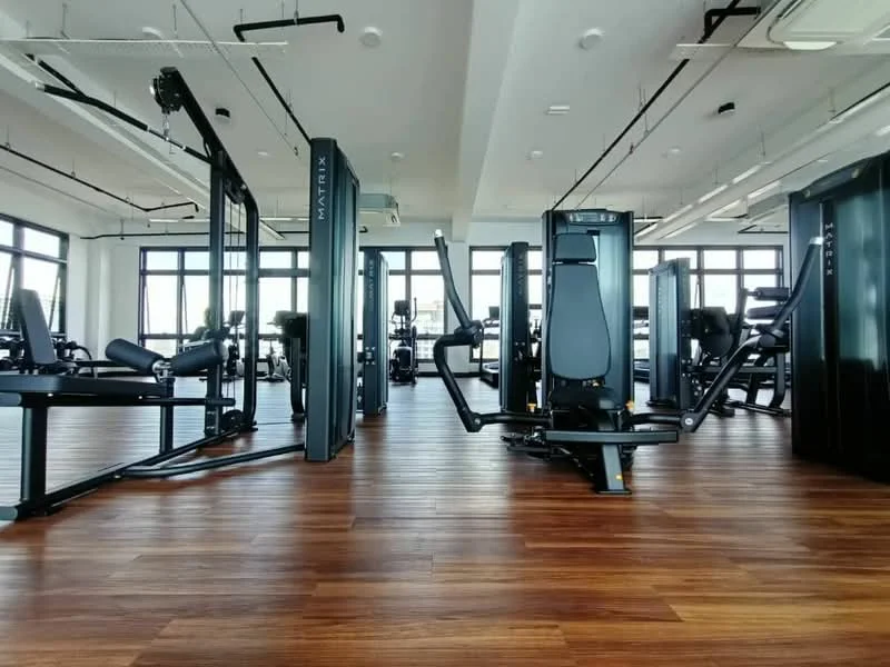 Gym features: wooden flooring, large windows with city view, various gym equipment, strength training machines, cardio machines, mirrored wall, ceiling-mounted air conditioning units, high ceilings, open layout; style: modern; view: city.