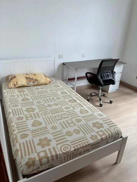 Bedroom features: single bed with patterned bedding, wooden bed frame, study desk with drawers, ergonomic office chair, laminate flooring, white painted walls, window with natural light, decorative pillow on bed; style: minimalist.