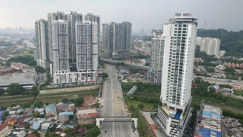 Exterior type: high-rise condominium; features: glass curtain wall, recessed balconies, surrounded by urban landscape, multiple high-rise buildings, main road in foreground, greenery along the road; style: modern.