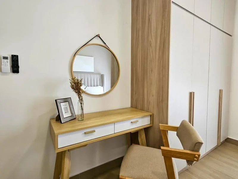 Room features: wooden desk with two drawers, round mirror, beige upholstered chair, white wall, light wood cabinetry, light wood flooring, decorative vase with dried flowers, framed artwork; style: modern.