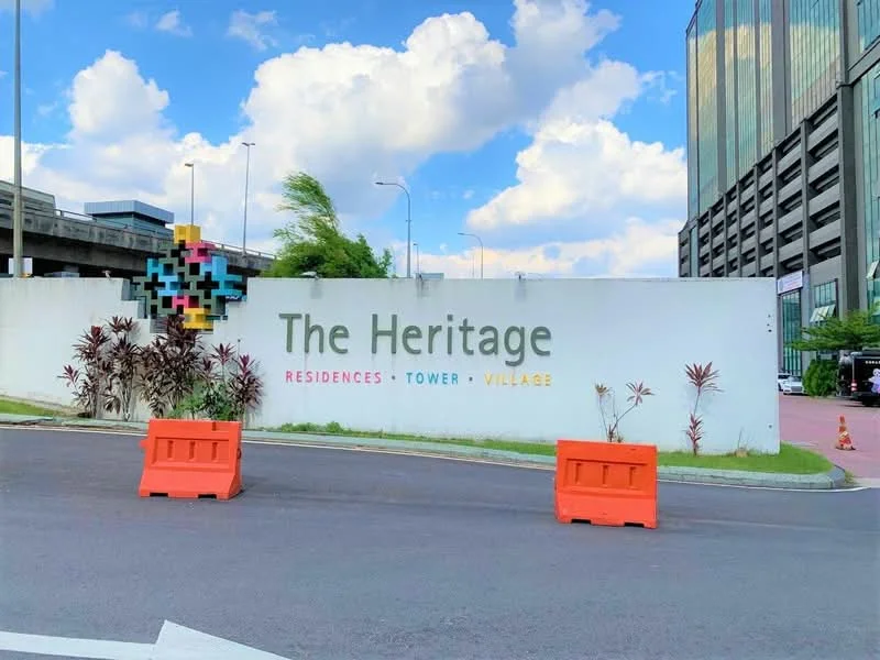 Exterior features: white painted facade, colorful decorative elements, landscaped area with plants, road access, traffic barriers, surrounding greenery, signage with the name 'The Heritage', cloudy sky; style: modern.