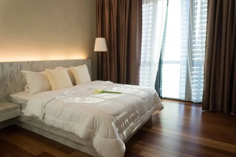 Bedroom features: king-size bed with white bedding, upholstered headboard, two white pillows, wooden flooring, floor-to-ceiling windows, sheer curtains, dark brown curtains, wall-mounted lamp, decorative flower on bed, light-colored accent wall; style: modern.