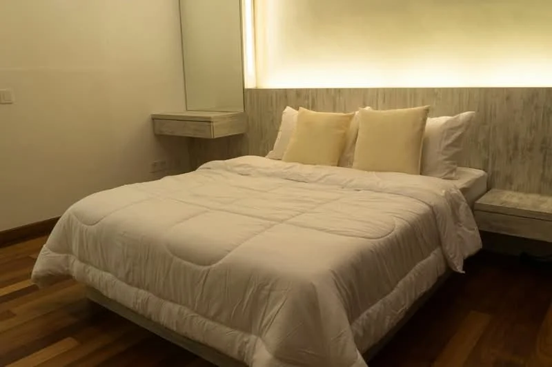 Bedroom features: king-size bed with white bedding, two beige decorative pillows, wooden flooring, wall-mounted bedside table, backlit wall feature, mirror above the bedside table, neutral wall color, soft ambient lighting; style: modern.