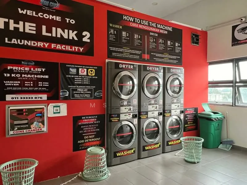 Laundry features: red painted walls, grey tiled flooring, coin-operated washing machines, coin-operated dryers, laundry baskets, price list on wall, instructions for machine use, detergent dispenser, green trash bin.
