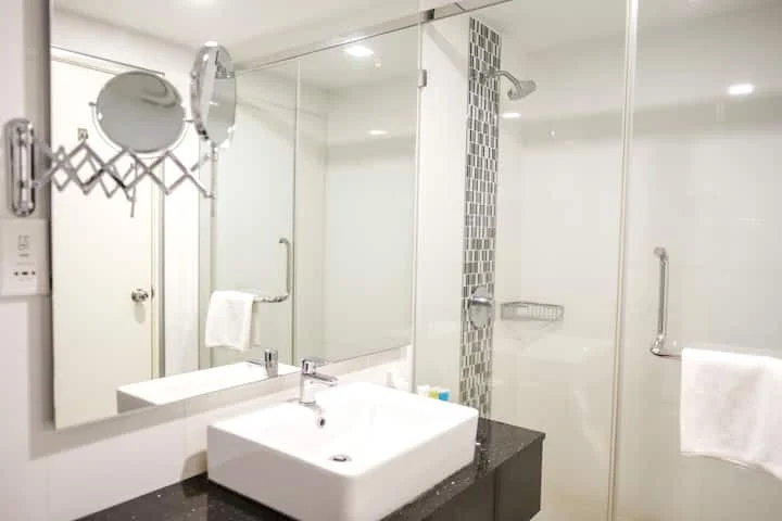 Bathroom features: white square sink, chrome basin mixer faucet, black countertop, large wall mirror, glass shower enclosure, rain shower head, tiled accent wall with mosaic pattern, white towels, wall-mounted towel rack, recessed lighting; style: modern.