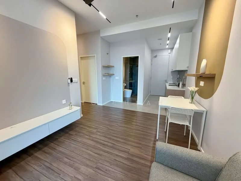 Living features: light grey accent wall, white painted walls, laminate flooring, recessed downlights, wall-mounted TV console, open shelving, dining table for two, grey fabric sofa, decorative vase with flowers, doorway to kitchen, doorway to bathroom; style: modern. Kitchen features: white cabinetry, stainless-steel sink, built-in oven, freestanding range oven, refrigerator, countertop with bar stools; style: modern. Bathroom features: one-piece toilet, shower with glass enclosure, vanity with mirror; style: modern.