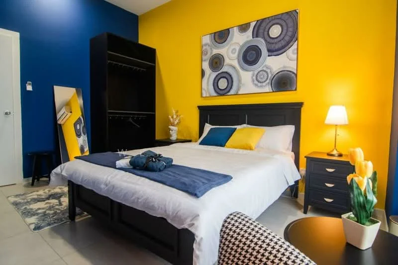 Bedroom features: king-size bed with white bedding, upholstered headboard, two decorative pillows in blue and yellow, nightstand with lamp, framed artwork with circular patterns, built-in wardrobe with open shelving, full-length mirror, area rug with geometric pattern, wall-mounted air conditioning unit, grey accent wall, yellow painted wall, black and white patterned chair; style: modern.