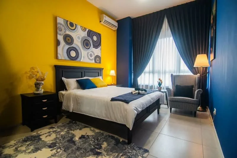 Bedroom features: queen-size bed with white bedding, upholstered headboard, two bedside lamps, black wooden nightstand, decorative artwork on wall, blue accent wall, yellow accent wall, floor-to-ceiling windows with sheer curtains, grey armchair, area rug with floral pattern, air conditioning unit, wall-mounted light fixture; style: modern.