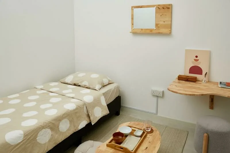 Bedroom features: single bed with polka dot bedding, wooden bedside table, decorative wall mirror, light-colored walls, textured area rug, wooden wall shelf, minimalist artwork, glass drinking cup, ceramic bowl, wooden tray; style: minimalist.