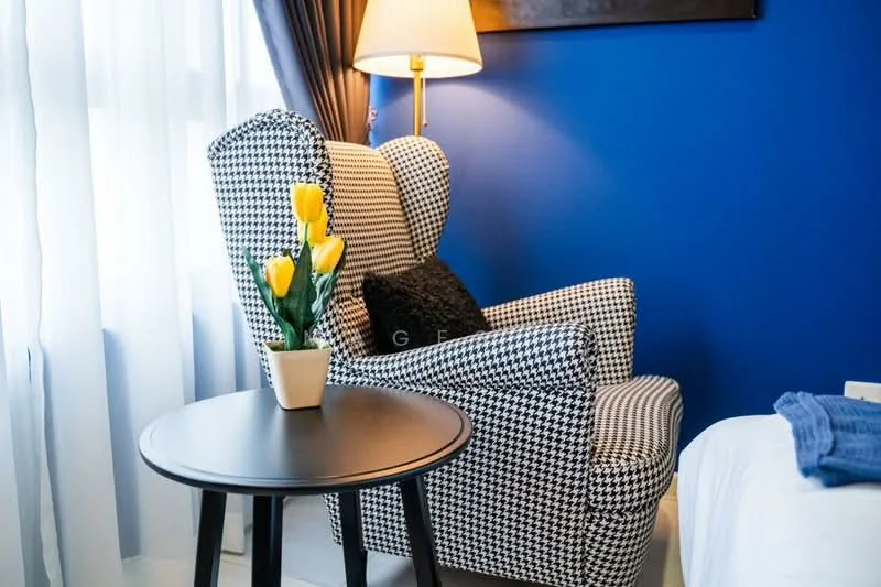 Room features: blue painted walls, checkered armchair in black and white, black side table, decorative plant in pot with yellow flowers, floor-to-ceiling window with sheer curtains, lamp with warm light; style: modern.