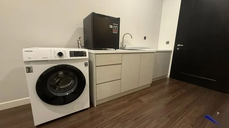 Laundry features: white front-load washing machine, black refrigerator, single-bowl stainless-steel sink, chrome pull-down faucet, light wood cabinetry, dark wood flooring, grey walls, countertop, wall-mounted electrical outlet; style: modern.