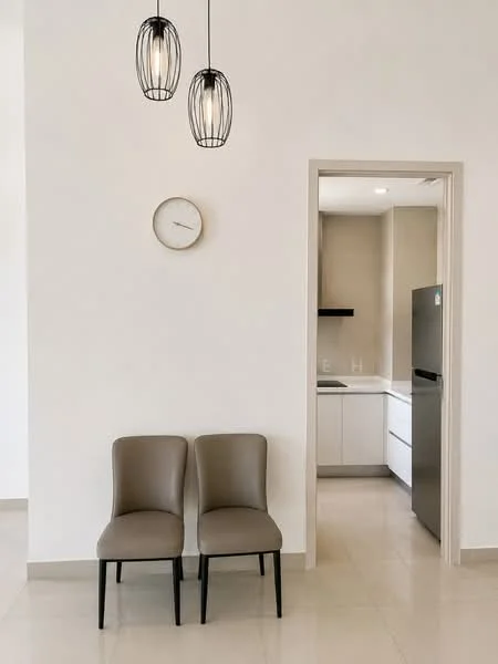 Room features: two beige dining chairs with black legs, white painted walls, light-colored tile flooring, pendant lighting fixtures, round wall clock, open doorway leading to kitchen, kitchen with white cabinetry, stainless-steel fridge visible in kitchen; style: modern.