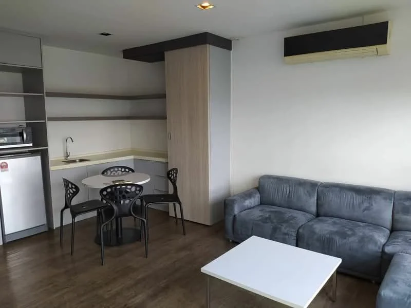 Living features: grey fabric sofa, white coffee table, round dining table, 4 black dining chairs, built-in shelving unit, kitchenette with sink, refrigerator, wood laminate flooring, wall-mounted air conditioning unit, ceiling lights; style: modern.