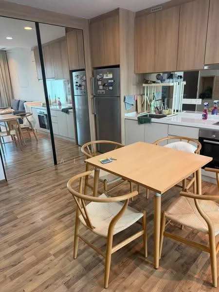 Dining features: light wood dining table, 4 wooden dining chairs with woven seats, laminate flooring, built-in cabinetry with wood finish, stainless-steel refrigerator, built-in oven, microwave, kitchen sink, countertop with white surface, mirror wall reflecting kitchen area, natural light from window, decorative items on countertop; style: modern.