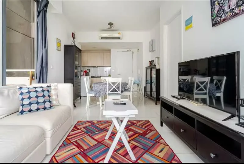 Living features: white leather sofa, decorative throw pillows with geometric pattern, area rug with colorful geometric pattern, coffee table, wall-mounted TV, TV console with storage, recessed ceiling lights, floor-to-ceiling windows, curtains, air conditioning unit, dining area visible in background, open-plan layout; style: modern. Dining features: 4-seater dining table, upholstered dining chairs, tablecloth, kitchen area visible in background; style: modern.