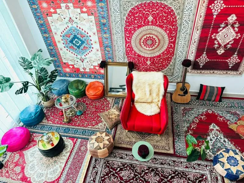 Room features: red accent chair, various patterned rugs, colorful floor cushions, decorative wall tapestries, indoor plants, wooden guitar, mirror, small round table, decorative bowls, faux fur throw; style: bohemian.