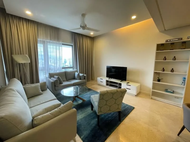 Living features: 3-seater fabric sofa in light grey, 2-seater fabric sofa in light grey, accent chair with patterned upholstery, glass coffee table, TV console in white, wall-mounted TV, built-in shelving unit, floor-to-ceiling windows with sheer curtains, recessed downlights, ceiling fan, marble flooring, area rug in blue, decorative items on shelves; style: modern; view: city.