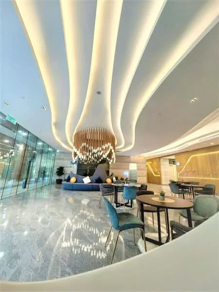 Room features: curved ceiling with recessed lighting, chandelier with hanging wooden beads, marble flooring, large glass windows, blue seating area with cushions, round dining tables, upholstered dining chairs in various colors, decorative plants, modern wall paneling with LED lighting; style: modern; view: city.