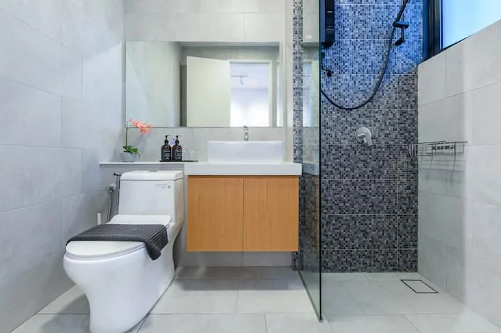 Bathroom features: single-sink vanity with wood cabinet, white countertop, framed mirror, one-piece toilet, walk-in shower with glass enclosure, rain shower head, handheld shower, tile cladding, grey mosaic tile accent wall, light grey ceramic tile flooring, chrome faucet, shower niche, towel rack; style: modern.