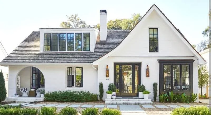 Exterior type: detached bungalow; features: white painted facade, pitched tile roof, black framed windows, arched entryway, two wall-mounted lanterns, manicured landscaping, stone-paved walkway, planters with greenery; style: modern.