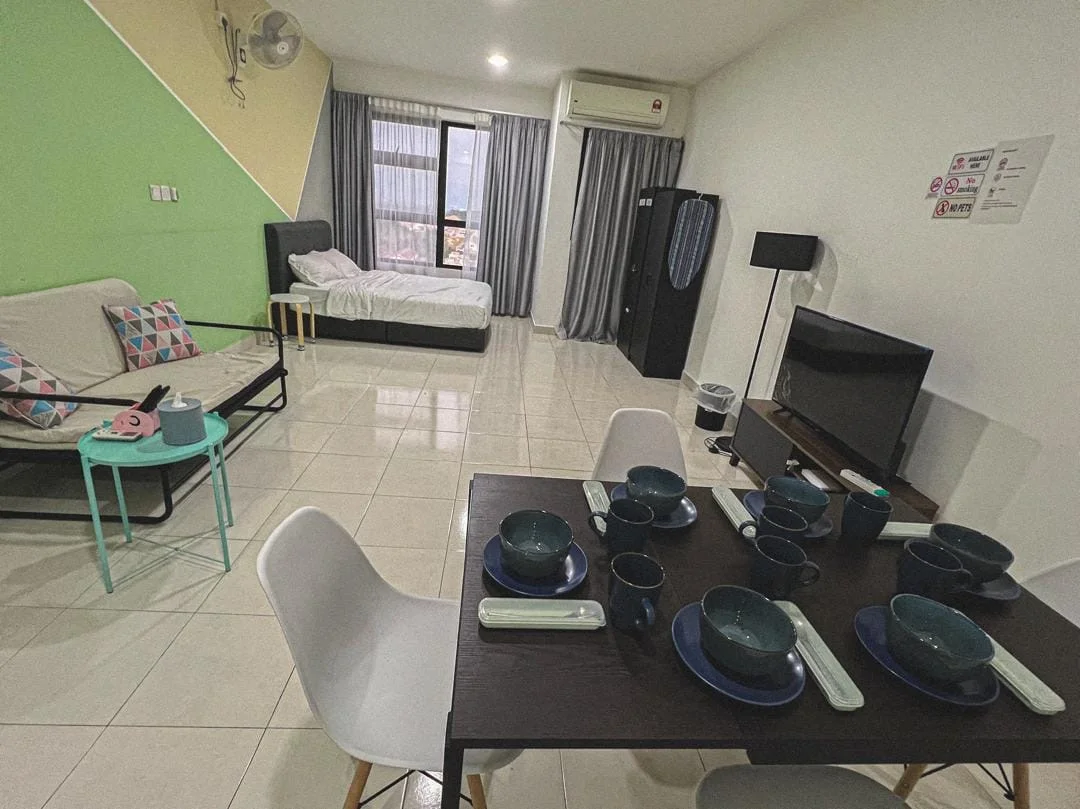 Living features: white painted walls, green accent wall, tile flooring, floor-to-ceiling windows, curtains, sofa bed with colorful cushions, black bed frame with white bedding, TV console, wall-mounted TV, dining table with 6 chairs, blue plates, blue bowls, cutlery, refrigerator, air conditioning unit; style: modern; view: city.