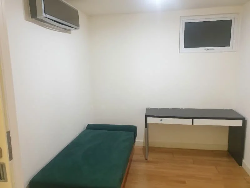 Bedroom features: single bed with green upholstery, wooden flooring, white painted walls, small window with black frame, wall-mounted split AC, desk with drawer in black and silver, minimalist decor; style: modern.