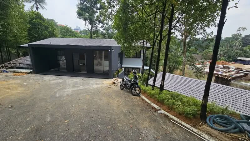 Exterior type: detached bungalow; features: flat roof, dark grey exterior walls, large glass windows, surrounded by trees, manicured landscaping, driveway with gravel surface, motorcycle parked in front, adjacent construction site, sloping terrain; style: modern.