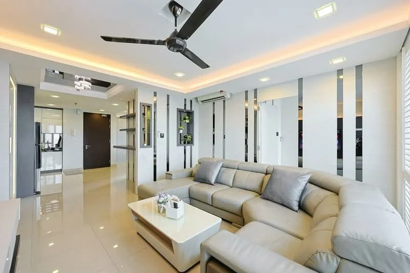 Living features: L-shape sectional sofa in light grey, coffee table with white surface, wall-mounted TV, recessed LED downlights, 5-blade ceiling fan with light, cream painted walls, glossy tile flooring, decorative wall with vertical mirrors, air conditioning unit, decorative plants on shelves; style: modern.
