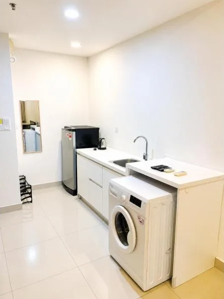 Laundry features: white painted walls, glossy white tile flooring, recessed ceiling downlights, single-bowl stainless-steel sink, chrome gooseneck faucet, white countertop, white base cabinets, compact laundry counter, front-load washing machine, single-door fridge, small wall mirror, built-in shelving niche, minimalist white finishes, narrow utility room layout; style: modern.
