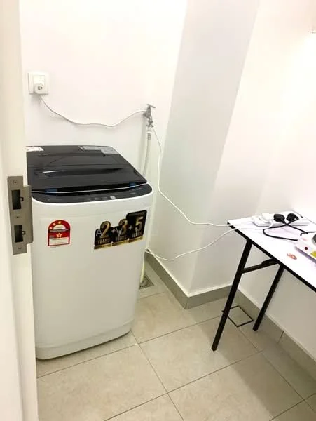 Laundry features: top-load washing machine in white, grey tiled flooring, white painted walls, small table with black legs, electrical outlet on wall, water supply connection, drainage outlet, power cord visible.