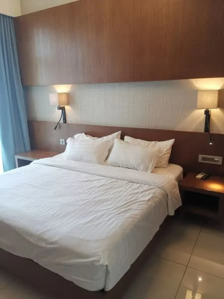 Bedroom features: king-size bed with white bedding, wooden headboard, two bedside lamps, light wood paneling on the wall, white painted walls, floor-to-ceiling window with blue curtains, white tile flooring, built-in bedside table; style: modern.