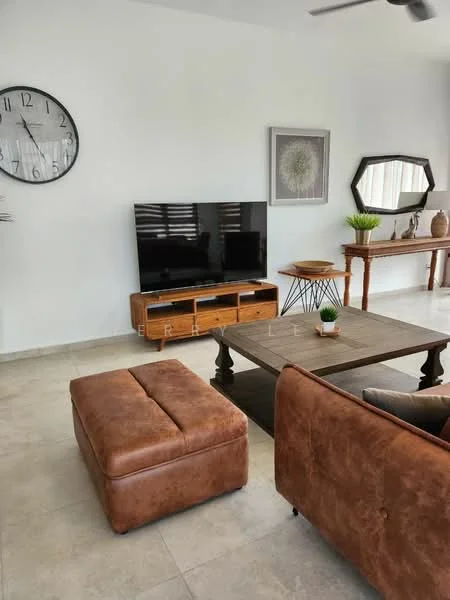 Living features: brown leather sofa, wooden coffee table, brown ottoman, wall-mounted flat-screen TV, wooden TV console, decorative wall mirror, framed artwork, clock on wall, plant in pot, window with blinds, light-colored tiled flooring, ceiling fan; style: modern.
