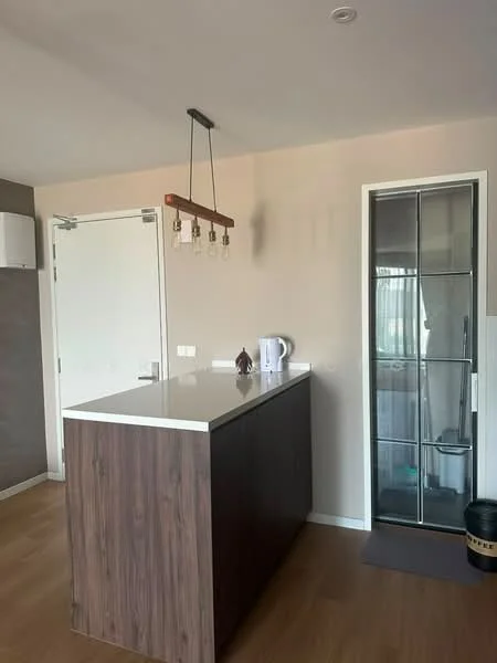Kitchen features: island countertop with dark wood finish, white quartz countertop, pendant lighting with exposed bulbs, built-in cabinet, stainless-steel kettle, glass shower enclosure, light wood flooring, neutral painted walls; style: modern.