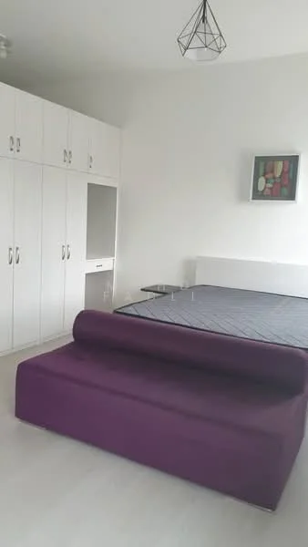 Bedroom features: white painted walls, light wood flooring, built-in wardrobe with white doors, purple sofa, queen-size bed frame without mattress, wall-mounted artwork, geometric pendant light; style: modern.