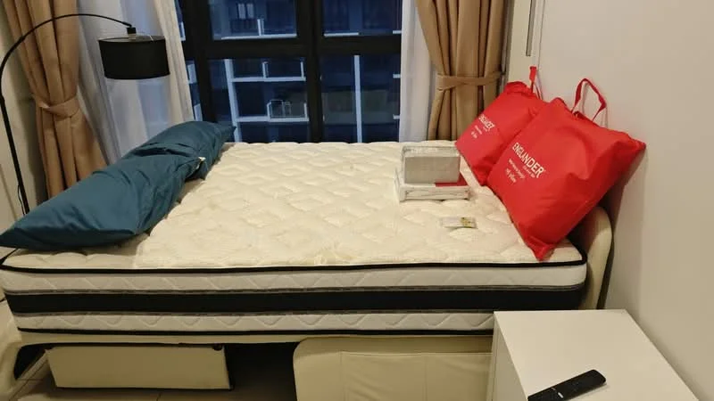Bedroom features: queen-size bed, white mattress, blue decorative pillow, red branded pillows, white bed frame, nightstand, table lamp, floor-to-ceiling windows, curtains, light-colored walls, tile flooring; style: modern; view: city.